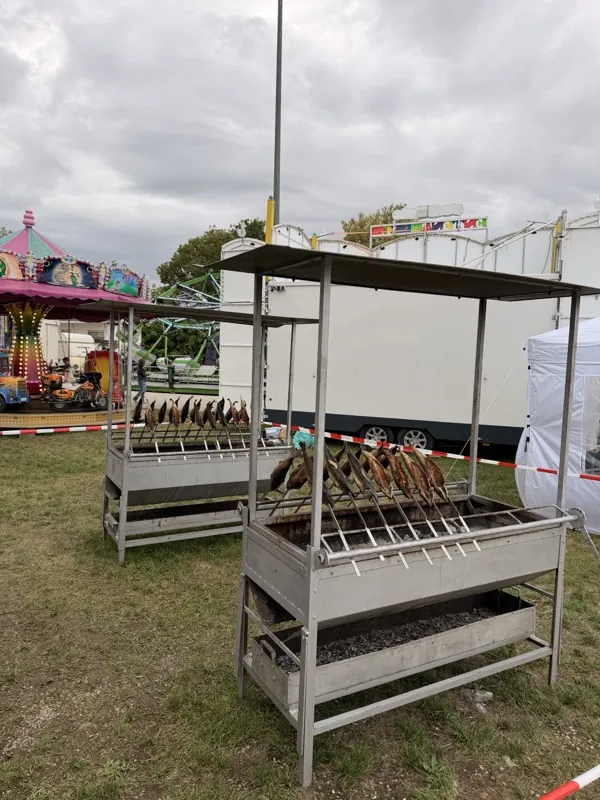 Grill voller Steckerlfische, im Hintergrund die Fahrgeschäfte des Volksfests